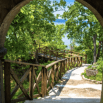 a path with wooden rails leading from a stone arch to a picturesque water body